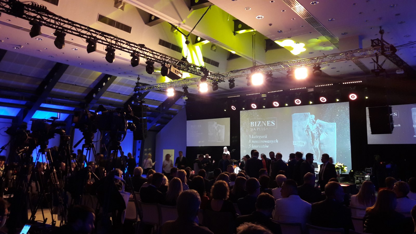 ludzie siedzą tłumnie zgromadzeni na sali. W głębi widać scenę