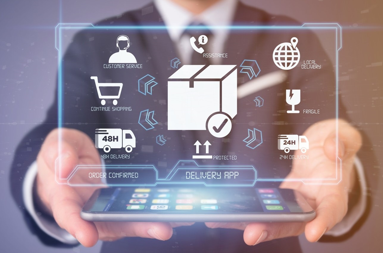 zbliżenie na mężczyznę w garniturze widzianego od pasa do szyi. Mężczyzna ma wyciągnięte obie dłonie do przodu, a w nich spoczywa smartfon. Na ekranie smartfona widać ikonki aplikacji. Nad smartfonem jest zaś nałożona cyfrowo abstrakcyjna grafika w formie hologromu, na której widać różne ikony i piktogramy symbolizujące handel elektroniczny