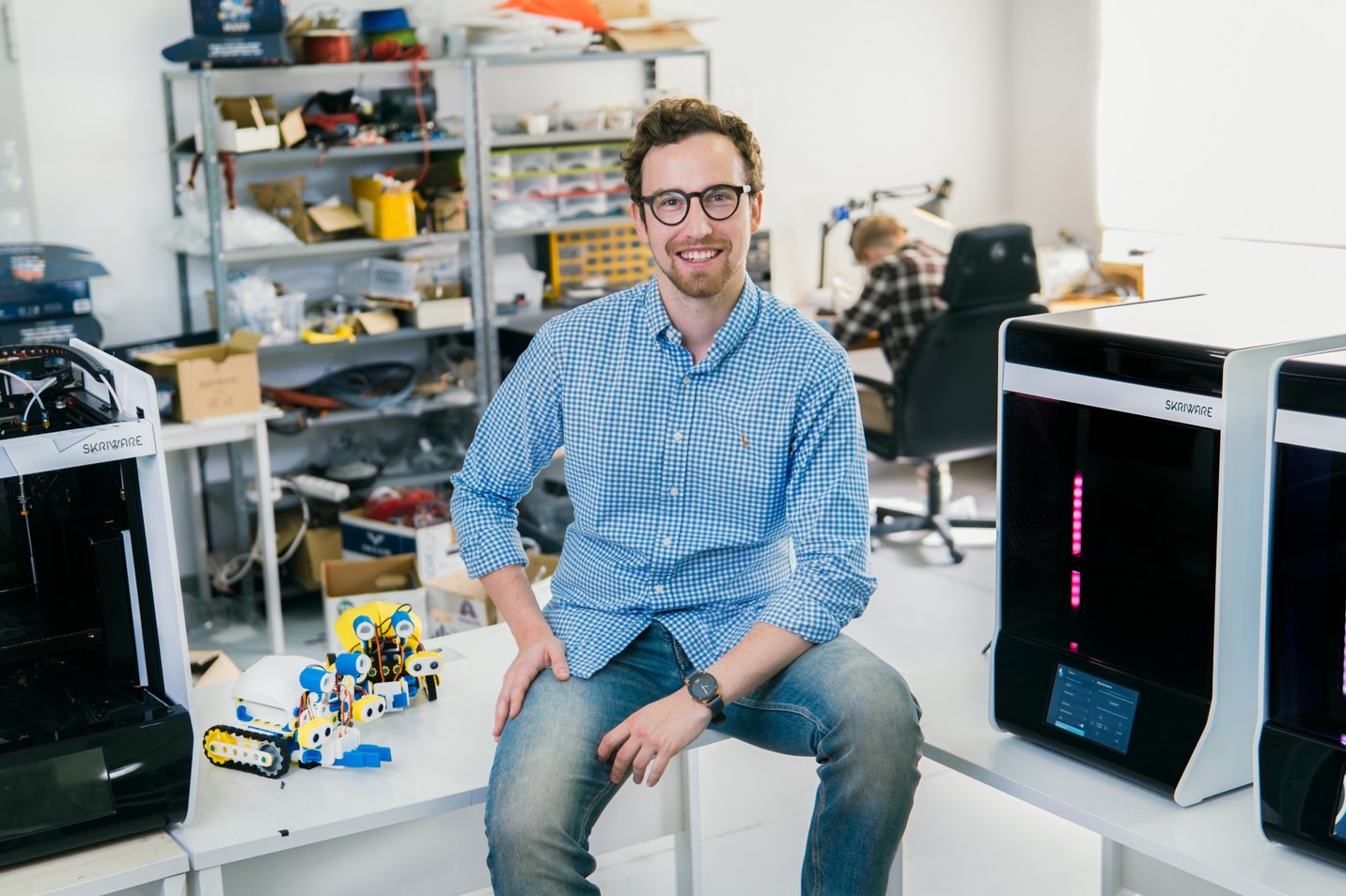 Młody mężczyzna w okularach siedzi na białym blacie. Obok niego, z lewej strony stoją dwa niewielkie roboty na gąsienicach, zaś obok nich drukarka 3D. Po przeciwnej stronie stoją z kolei dwie drukarki 3D. W tle widać metalowe regały oraz biurko, przy którym ktoś siedzi.