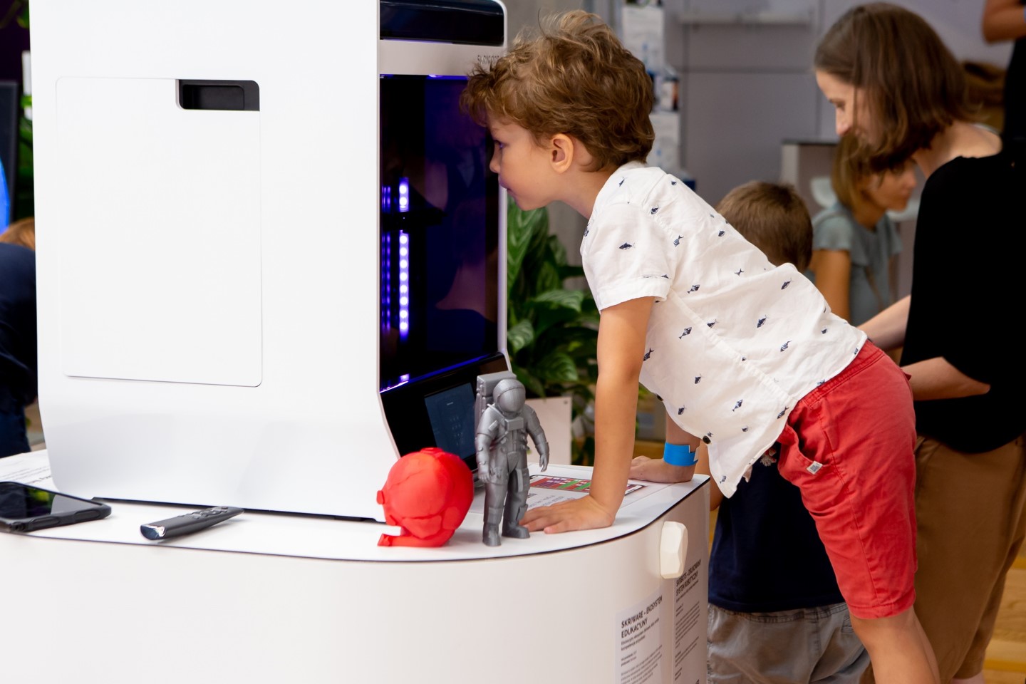 Chłopiec zagląda przez szybę do wnętrza obudowy drukarki 3D. Obudowa drukarki w białym kolorze. Jej szklany front tworzą ciemne szyby. W dolnej części znajduje się panel sterowania drukarki z wyświetlaczem LCD. Obok drukarki stoją dwa wydrukowane modele 3D w niewielkim rozmiarze – model anatomiczny serca w kolorze czerwonym oraz figurka astronauty w kolorze srebrnym. W głębi zdjęcia widać innego chłopca i dorosłą kobietę.