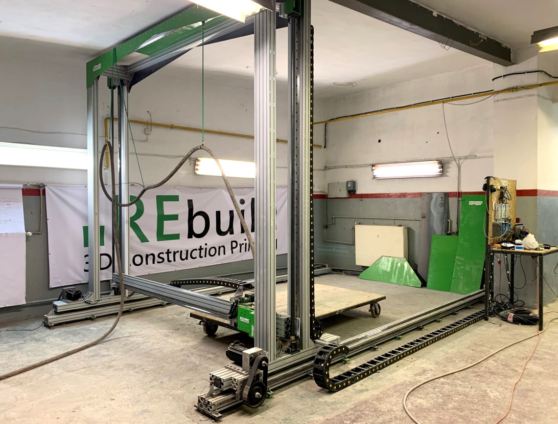 Zdjęcie przedstawia hale przemysłową, w której znajduje się metalowa konstrukcja, jej rama jest przytwierdzona do sufitu oraz ściany. Obok leżących na podłodze ramion ramy widać gąsienicę. W środku ramy widać zaś drewnianą płytę, do której od spodu przytwierdzone są koła. 