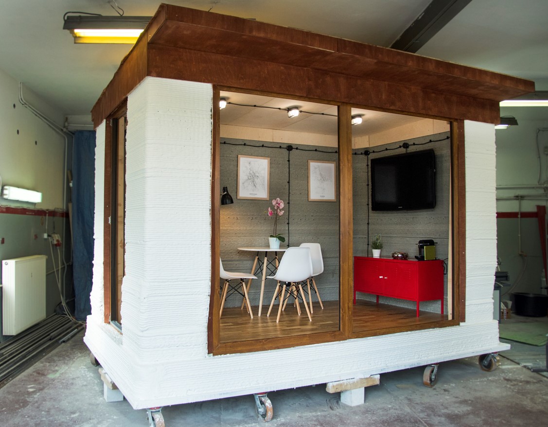 Widok na niewielkich wymiarów betonowy domek z centralnie umieszczonym oknem sięgającym od sufitu do podłogi. W środku widać stół, krzesła, komodę oraz wiszące na ścianie telewizor i obrazki.