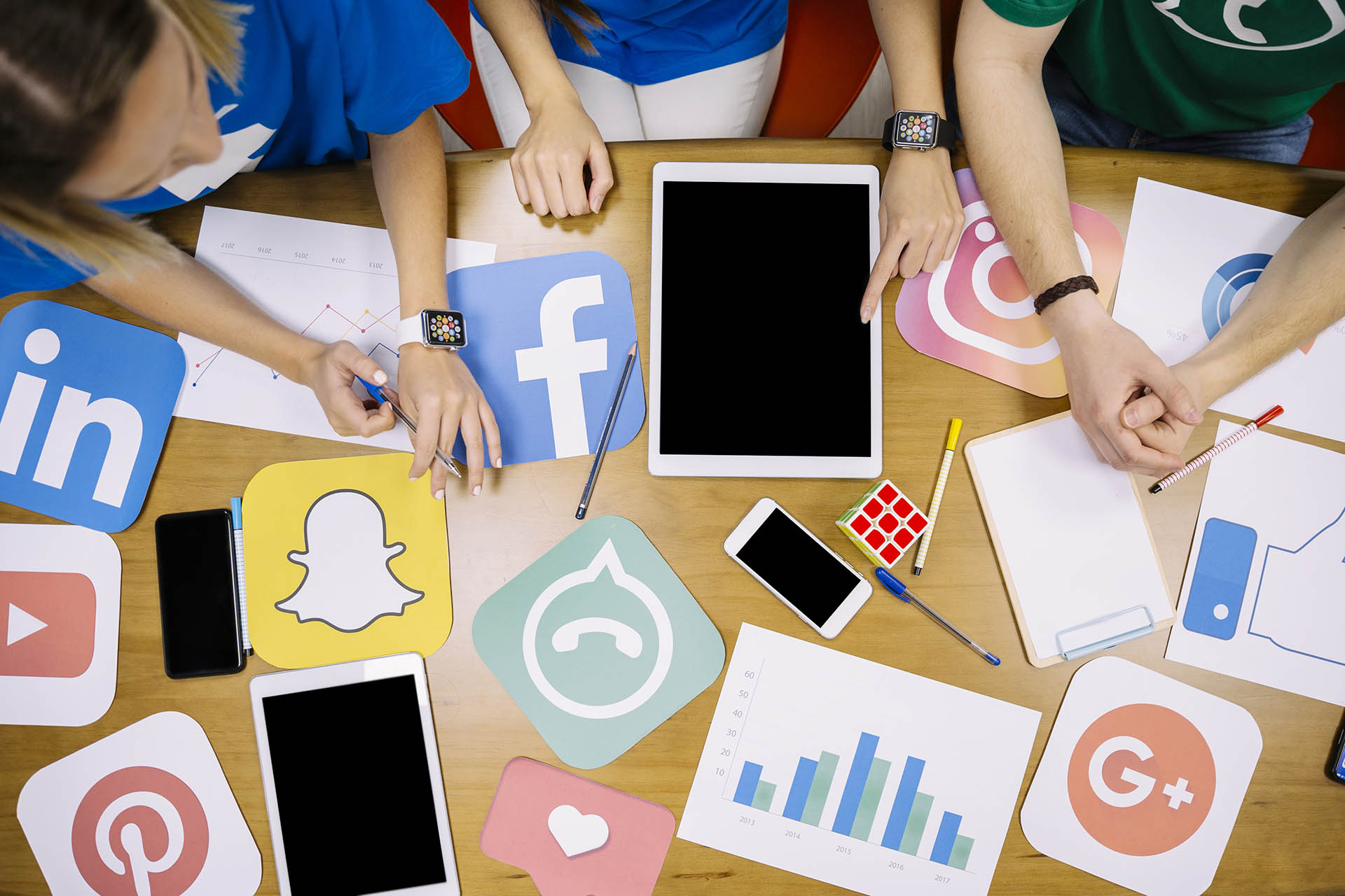 widok od góry na stół, przy którym siedzi trójka młodych ludzi, dwie kobiety i jeden mężczyzna. Na stole leżą tablety, kartki oraz logotypy ze znakami 