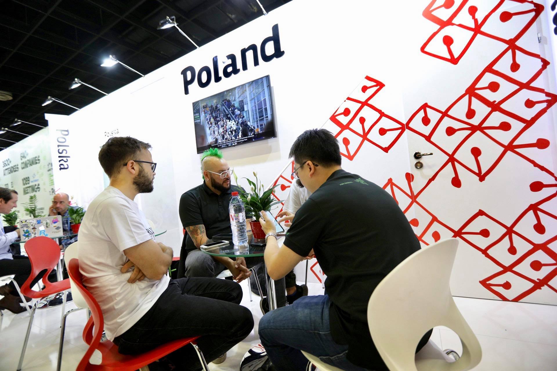 Cztery osoby siedzące przy stoliku. W tle biała ściana z monitorem i dużym napisem Poland